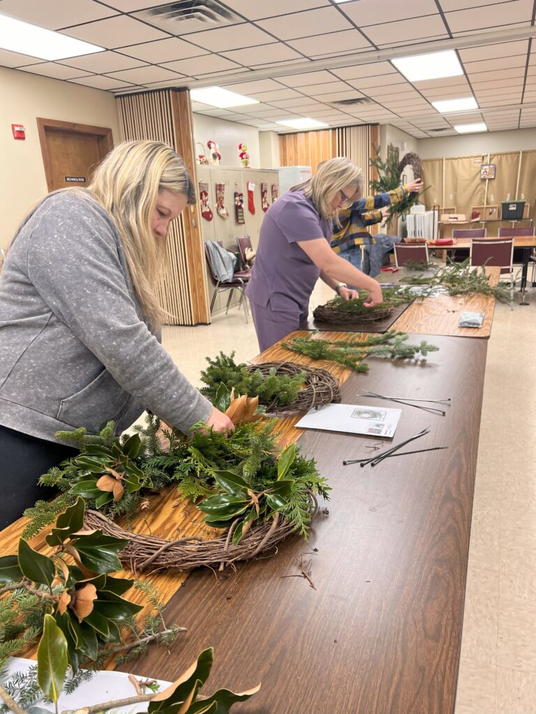 Women work, creating wreaths on a table.